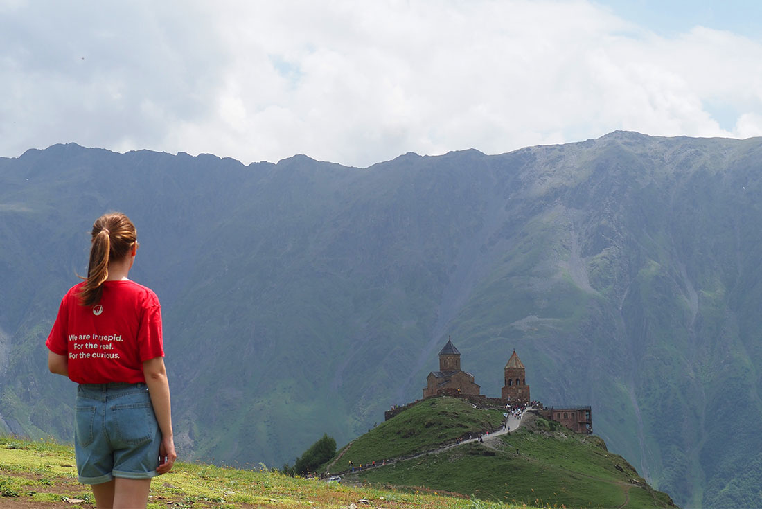 Intrepid leader stands at the crest of a hill looking out at the Gergeti Trinity Church with Mount Kazbek behind it in Georgia