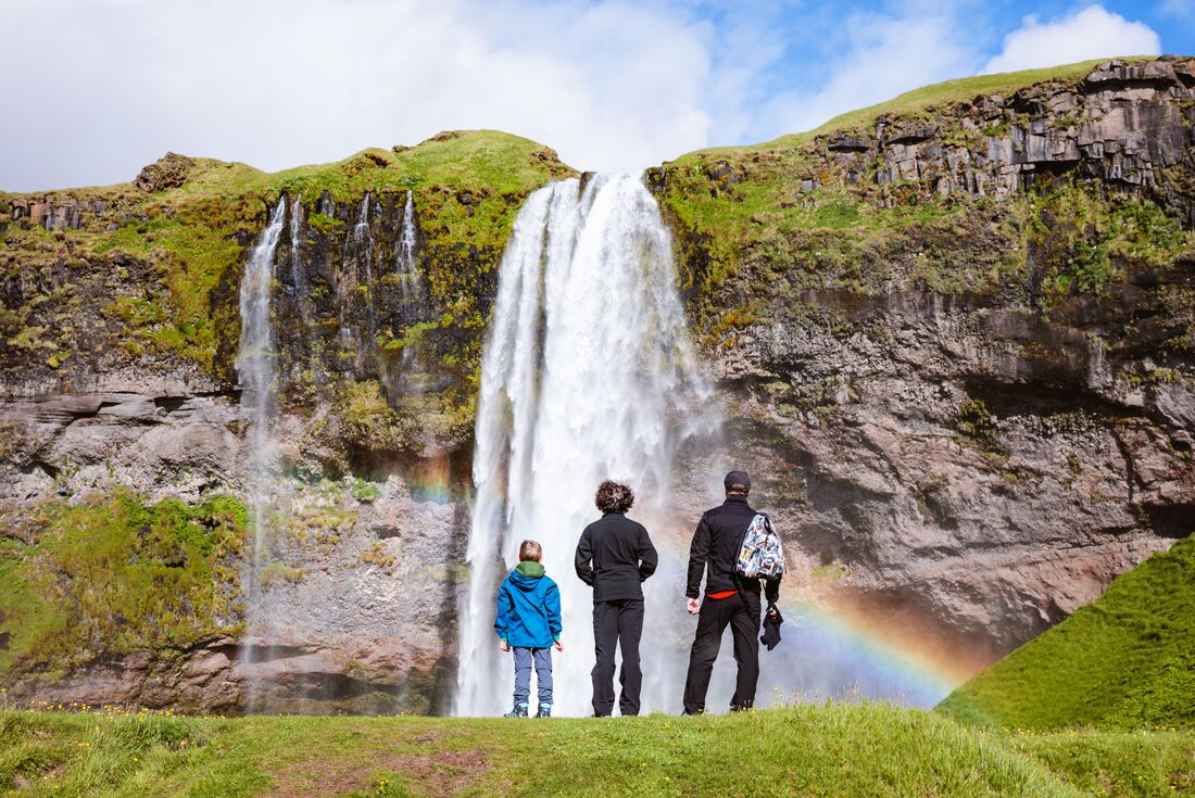 Family of travellers look at Seljalandsfoss waterfall as a rainbow forms in the mist in the Golden Circle of Iceland