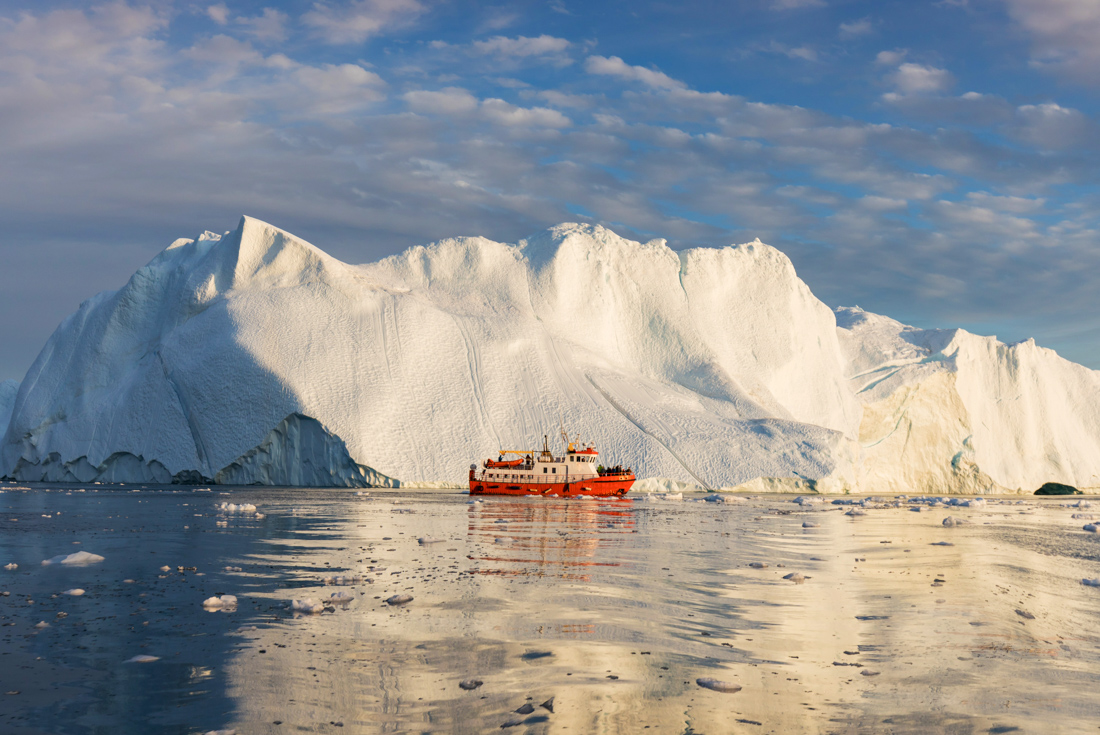 Small orange and white ship Disko Bay ferry overshadowed by huge iceberg in Disko Bay Greenland