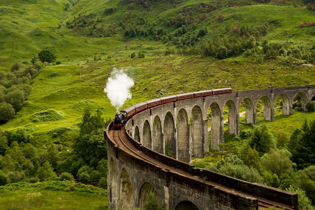 Aerial view of the Glenfinnan Viaduct with a steam engine train on it, winding above rolling green hills in Scotland, United Kingdom