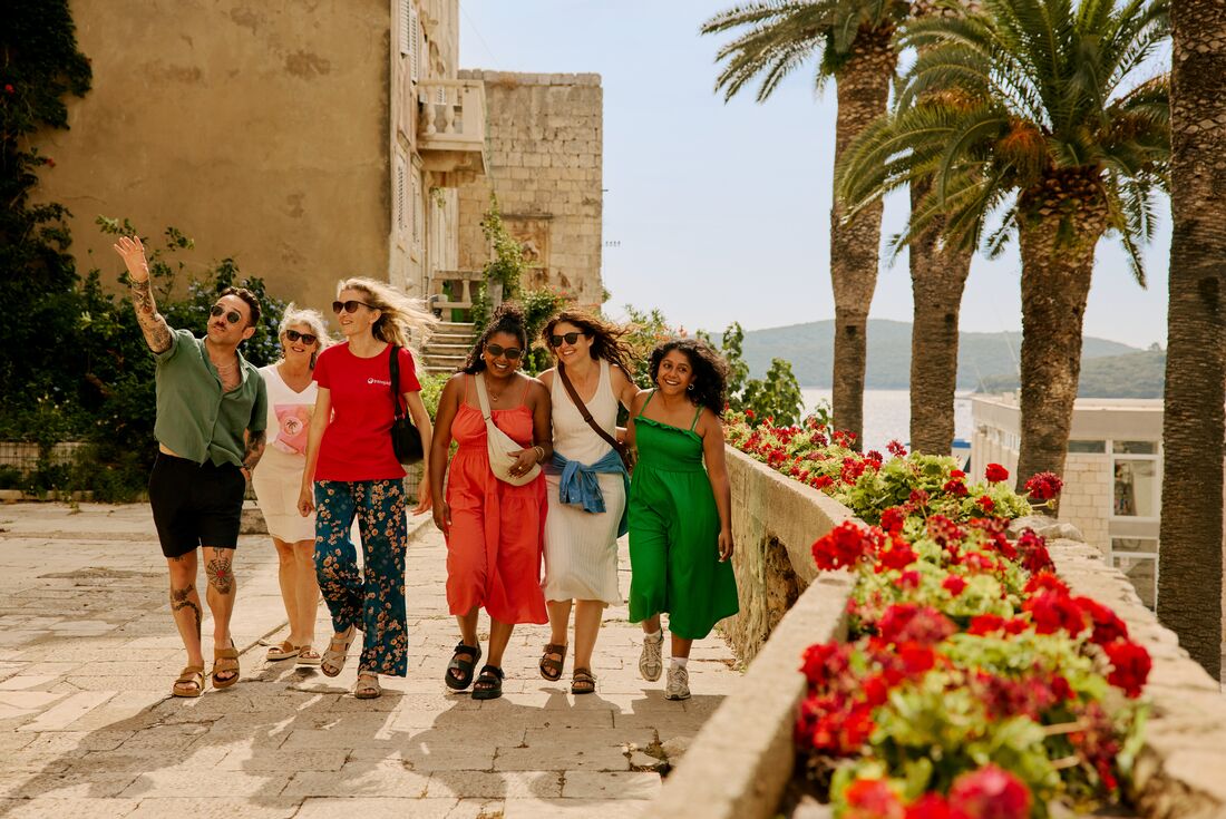 Travellers enjoying a walk with Leader in Korcula, lined with red flowers, palm trees in the background and mountainous landscape, Croatia