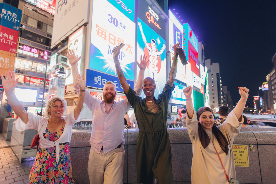 Intrepid travellers throw their hands up surrounded by screens and lights in Dotombori