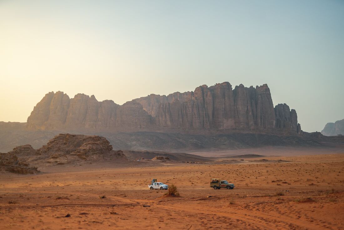 Spire mountain range in distance with two trucks driving past in the distance in an orange sand desert 