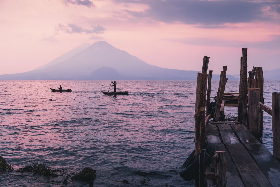 Locals on boats paddle across the reflective surface of Lake Atitlan at sunrise with volcano beyo9nd